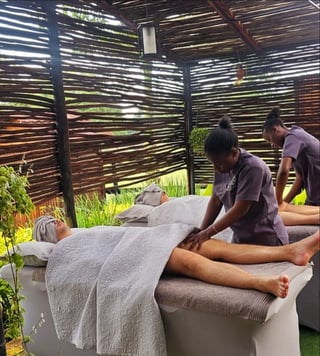 Two massage therapists providing treatment to a client in an outdoor spa setting with wooden lattice screens and potted plants
